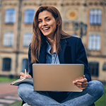 uśmiechnięta studentka studentka siedząca po turecku z laptopem i notatnikiem na kolanach