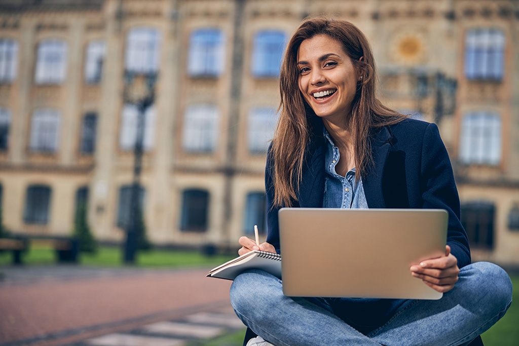 uśmiechnięta studentka studentka siedząca po turecku z laptopem i notatnikiem na kolanach